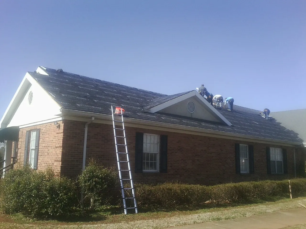 Crew tearing shingles off a brick ranch roof with a ladder set — Metal Roof Repair work by our crew in Ridgeway