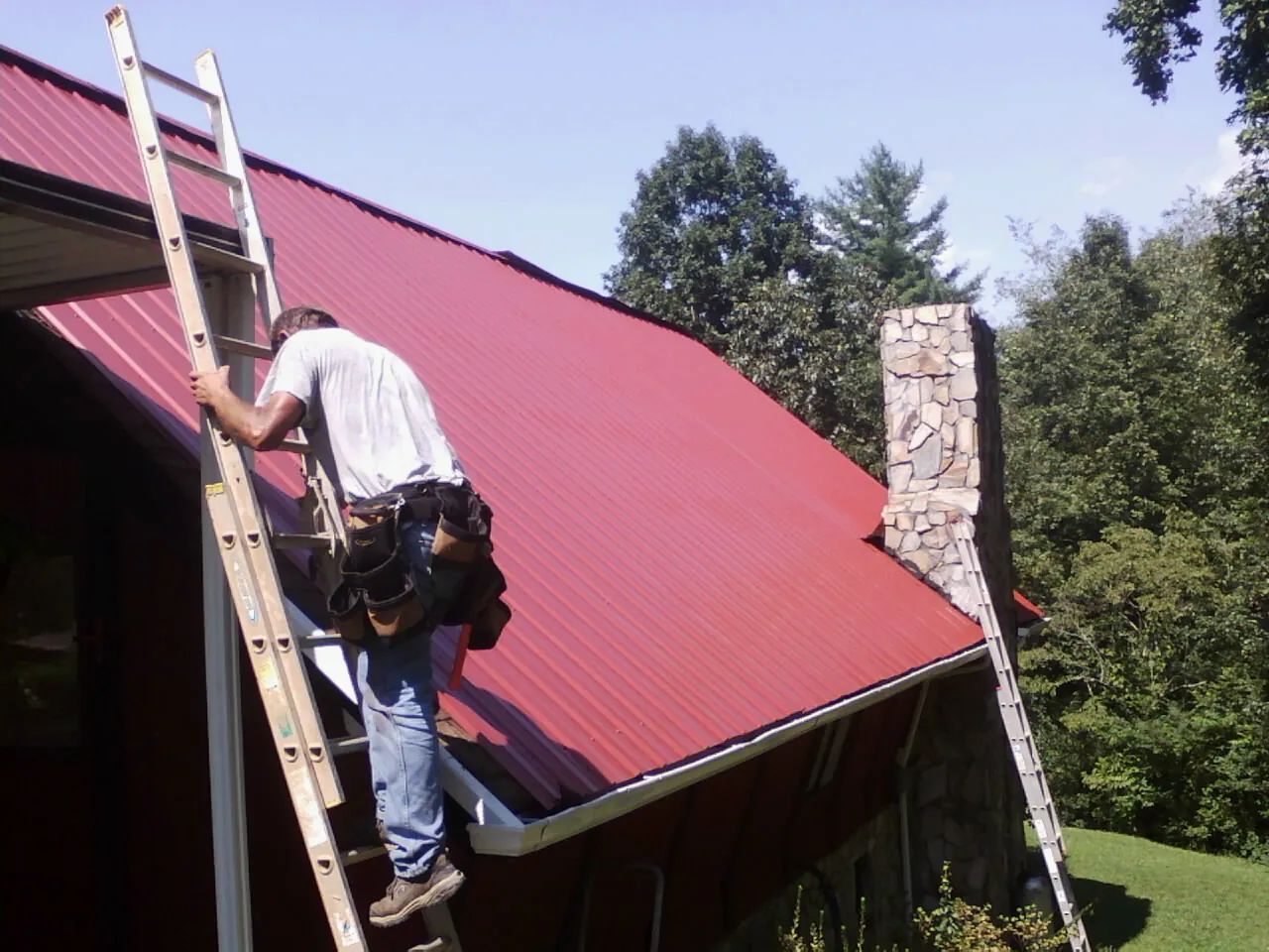 Roofer on a ladder beside a red metal roof and stone chimney — Soffit & Fascia Repair by Roofing Brothers in Ridgeway
